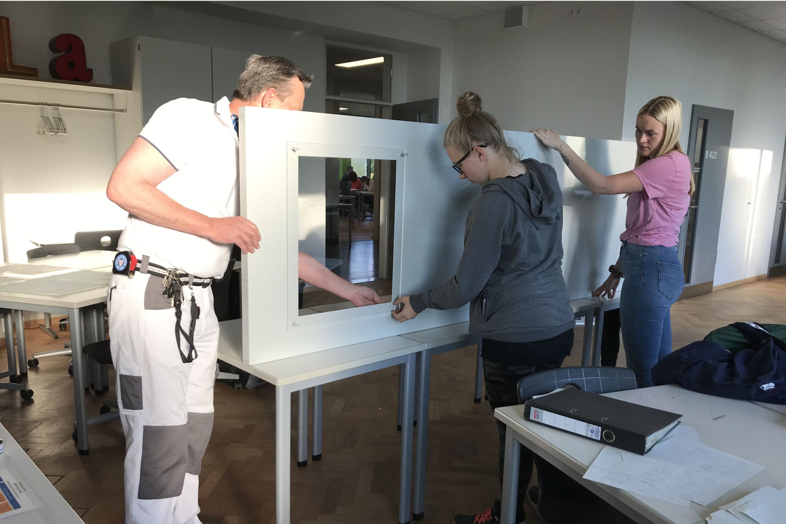 Zwei Schülerinnen und ein Lehrer haben eine Tür auf einem Tisch hochkant auf die Seite gestellt. Keine Ahnung, was da gemacht wird.
