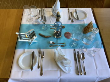 Festlich eingedeckter Tisch mit Strandbezug, Spielzeugliegestuhl und Muscheln.