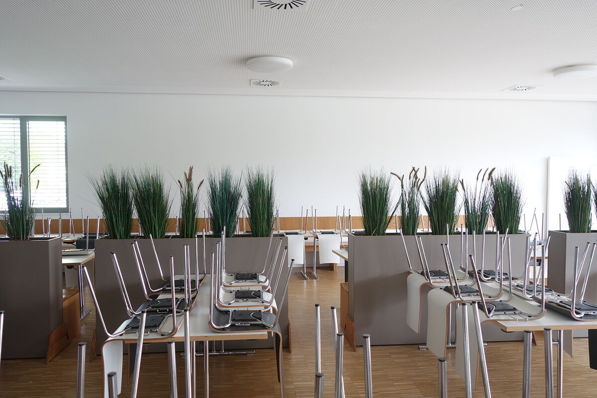 Leere Mensa mit hochgestellten Stühlen und vielen grasartigen Pflanzen in Pflanzkübeln.