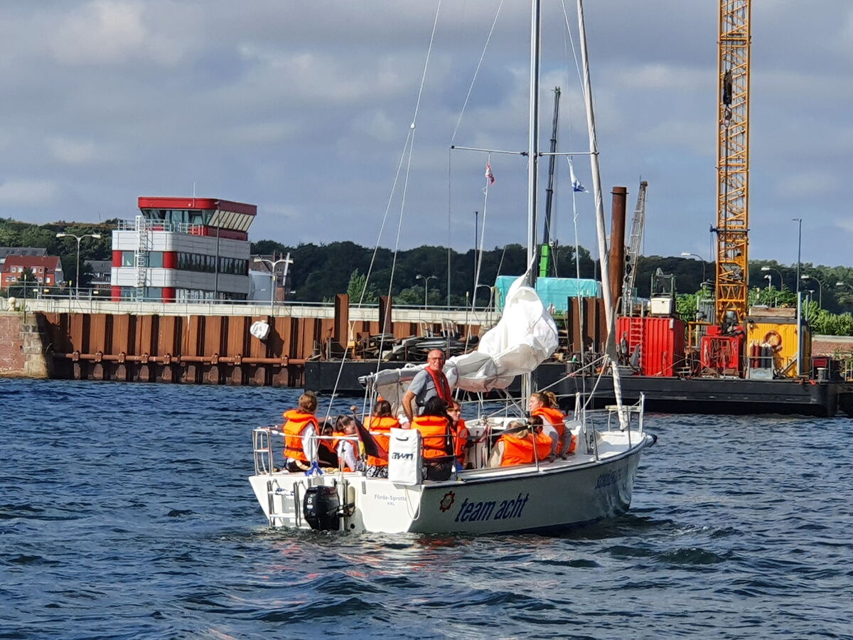 Gerefftes Segl und Besatzung mit Schwimmwesten vor einer Kaimauer.