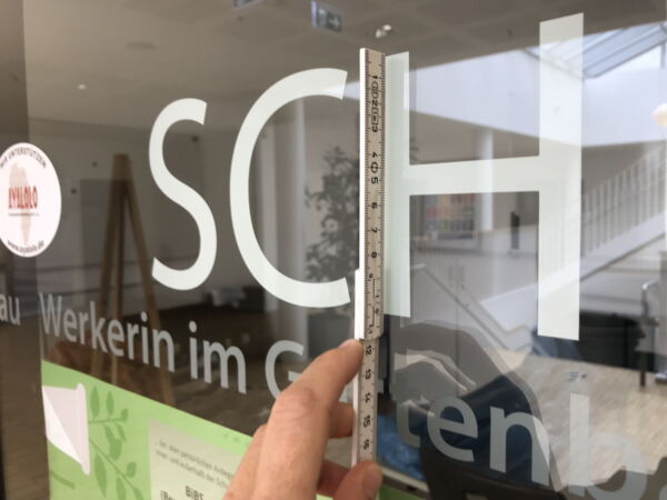 Mittels eines Zollstocks wird an einer Glasscheibe die Höhe des Buchstabens H gemessen.