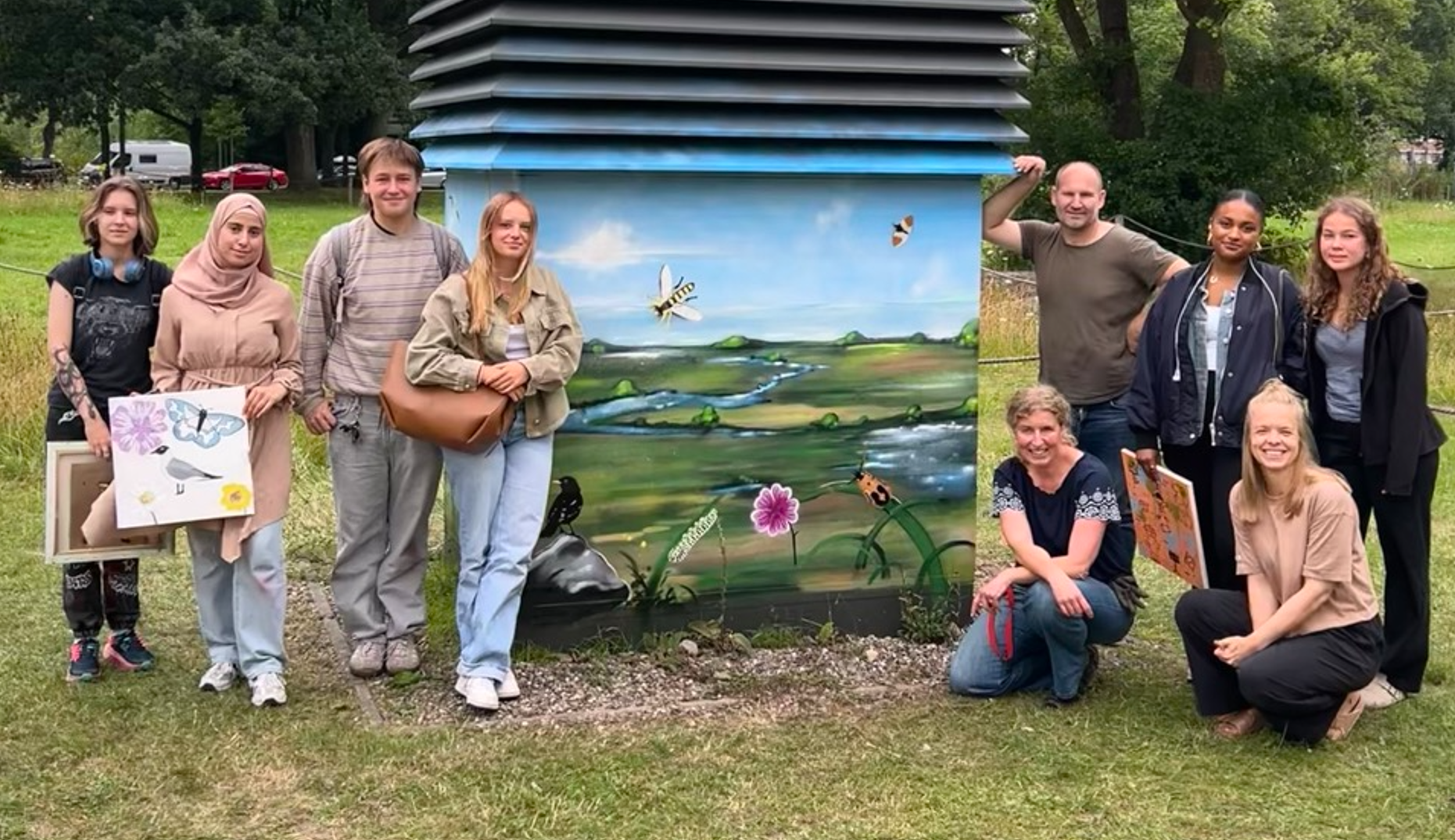 Die Schülergruppe vor der fertigen Enttlüftungsanlage, die mit Landschaft, Insekten und Wasserflächen besprüht ist.