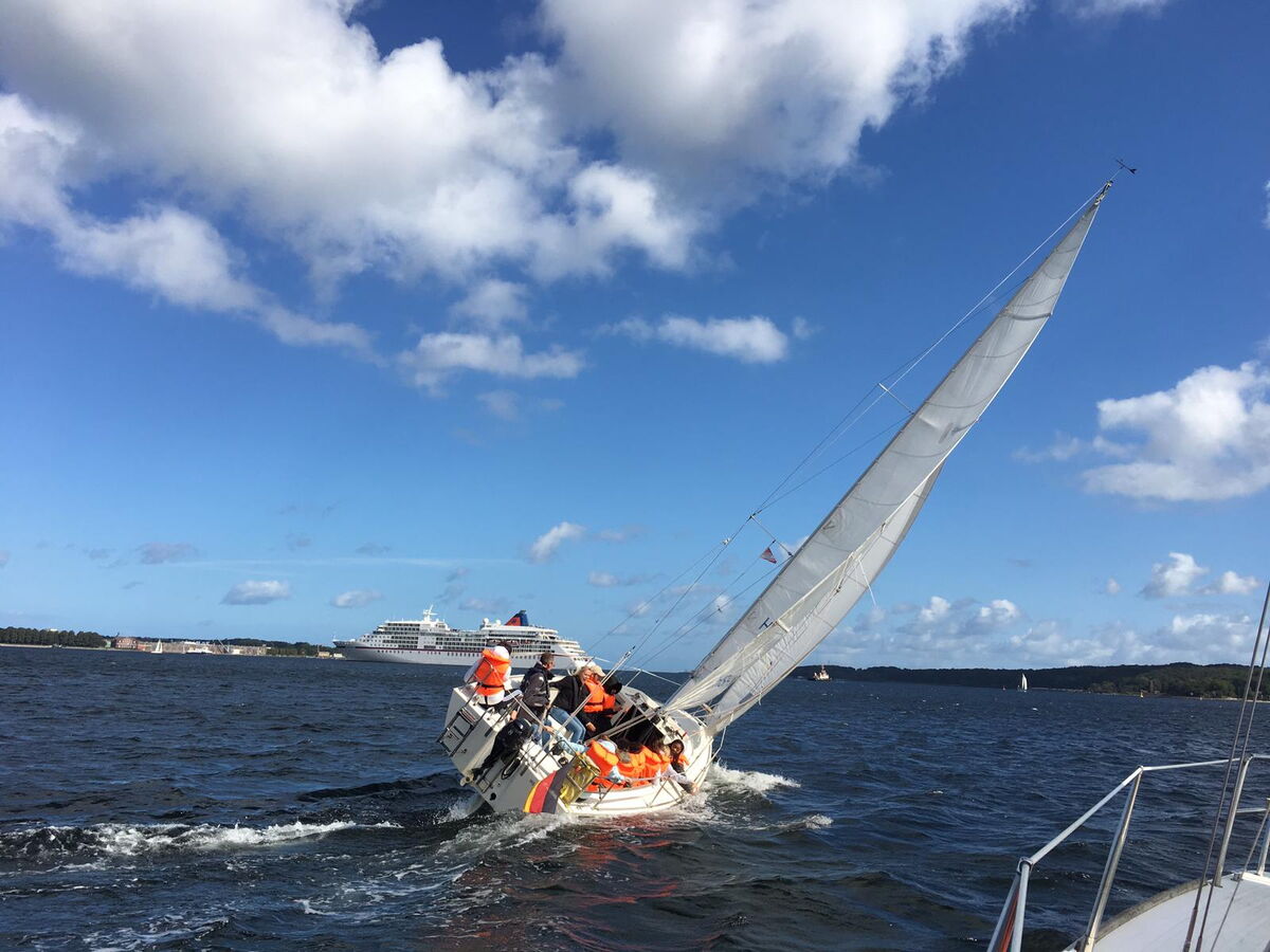 Schräg segelndes Boot auf der Kieler Förde mit einem Kreuzfahrtschiff im Hintergrund, blauer Himmerl und wenig Wolken.