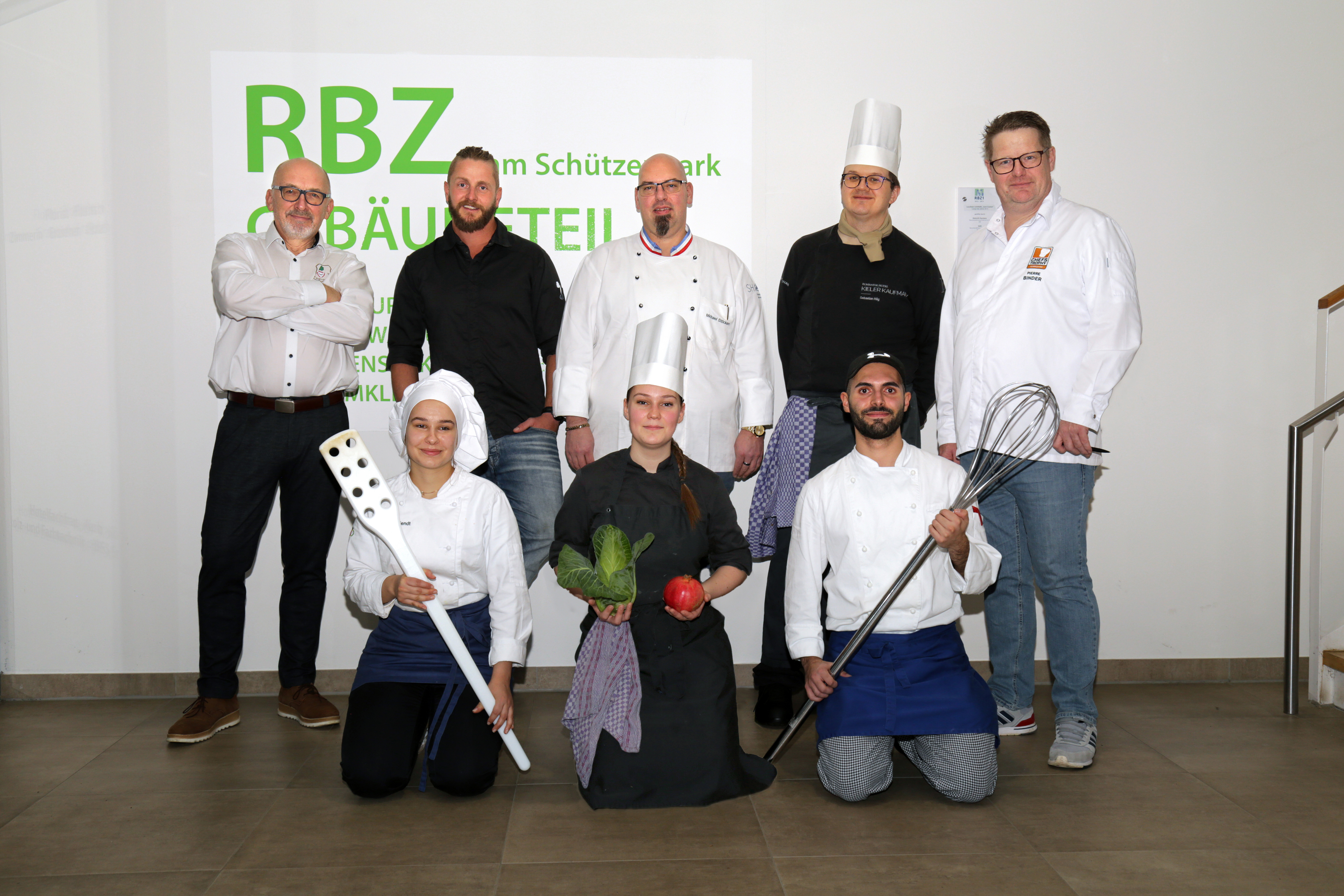 Köchinnen und Köche, zweireihig stehend und knieend, dabei übergroßes Besteck haltend.