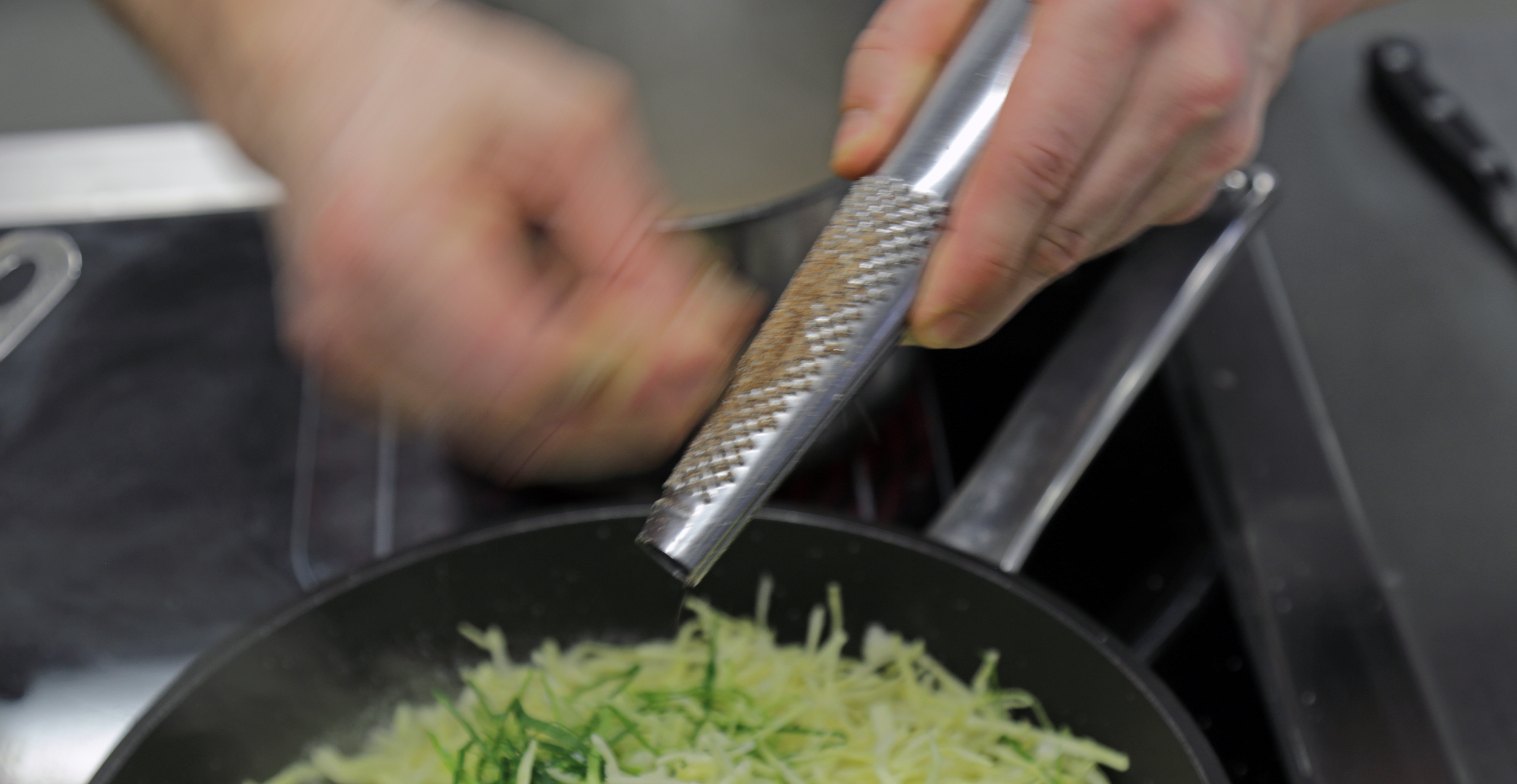 Ein Nahrungsmittel wird mit einer Raspel in eine Pfanne gerieben.