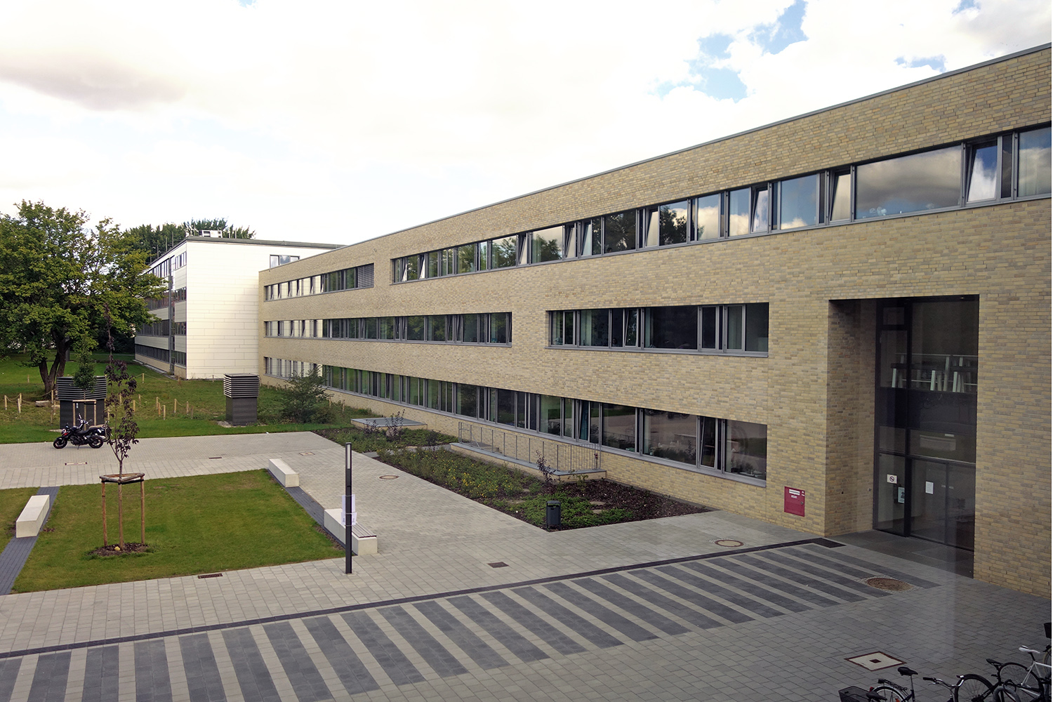 Zugang zum Eingang Westring mit Beeten, Rasenflächen, Pflasterung und abgestellten Fahrrädern.